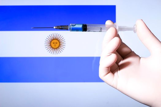 Hand in surgery glove holding syringe with covid vaccine. Argentina's flag in the background