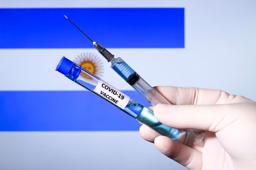 Hand in surgery glove holding syringe with covid vaccine. Argentina's flag in the background