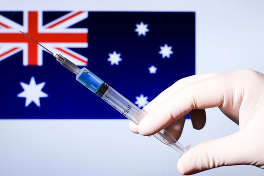 Hand in surgery glove holding syringe with covid vaccine. Australian flag in the background