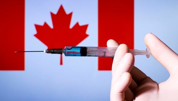 Hand in surgery glove holding syringe with covid vaccine. Canadian flag in the background