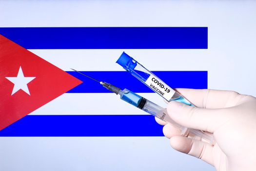 Hand in surgery glove holding syringe with covid vaccine. Cuban flag in the background