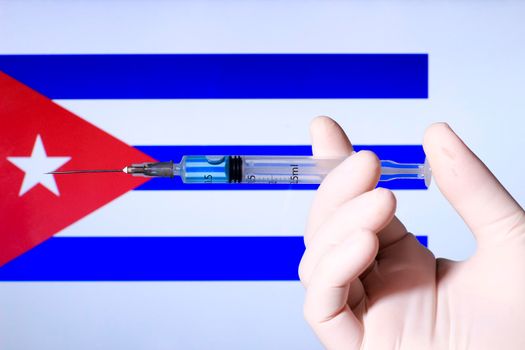Hand in surgery glove holding syringe with covid vaccine. Cuban flag in the background