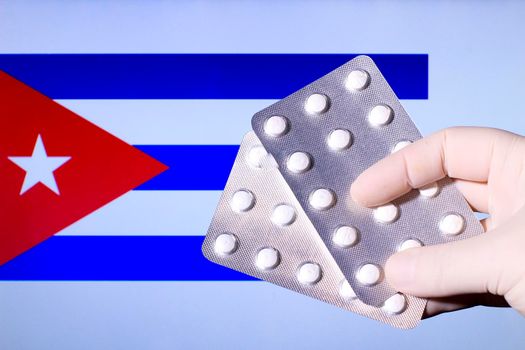 Hand in surgical glove holding pill blisters. Cuban flag in the background