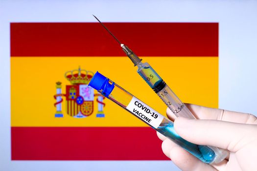 Hand in surgery glove holding syringe with covid vaccine. Spanish flag in the background