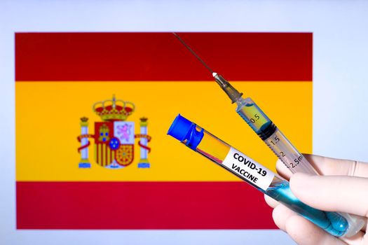 Hand in surgery glove holding syringe with covid vaccine. Spanish flag in the background