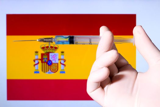 Hand in surgery glove holding syringe with covid vaccine. Spanish flag in the background