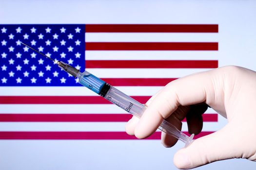 Hand in surgery glove holding syringe with covid vaccine. U.S. flag in the background
