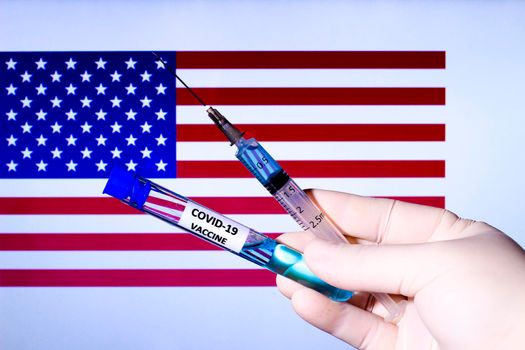 Hand in surgery glove holding syringe with covid vaccine. U.S. flag in the background