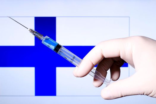Hand in surgery glove holding syringe with covid vaccine. Finnish flag in the background