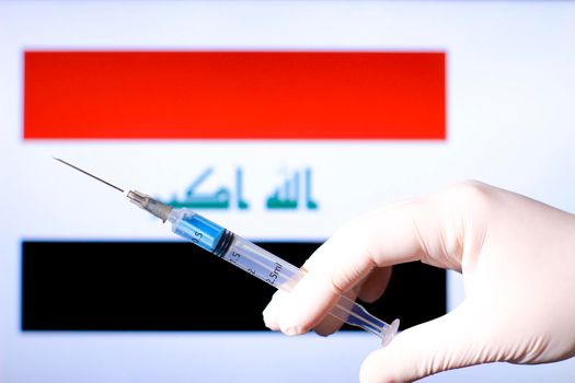 Hand in surgery glove holding syringe with covid vaccine. Iraqi flag in the background
