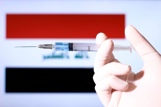 Hand in surgery glove holding syringe with covid vaccine. Iraqi flag in the background