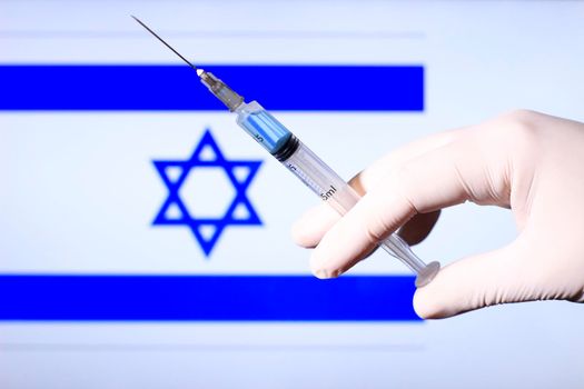 Hand in surgery glove holding syringe with covid vaccine. Israeli flag in the background