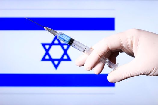 Hand in surgery glove holding syringe with covid vaccine. Israeli flag in the background