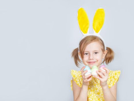 Portrait of a little cute Caucasian smiling girl with bunny ears and decorations for Easter in hands on a gray background. Easter background with place to insert text.