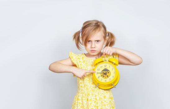 A Little Caucasian girl holds an alarm clock in her hand and looks angrily at the camera and points finger at the clock, advertisement posing against studio wall. Advertising concept