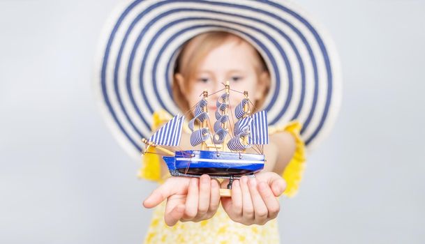 A Little Caucasian girl in a wide-brimmed hat holds out a toy boat forward. Focus on the foreground. The concept of summer vacation, travel, opening of the beach season..