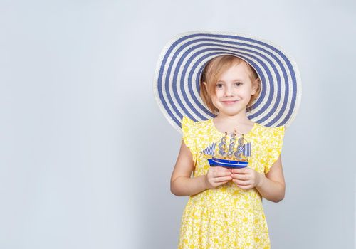 A Little Caucasian girl in a wide-brimmed hat holds a toy boat in her hands. The concept of summer vacation, travel, opening of the beach season.