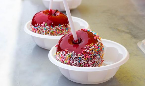 A red caramelized apple on a stick, covered in hard red sugar, standing in a plastic plate to serve as a festive treat. The concept of Easter, Maslenitsa,Valentine's Day.