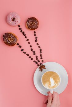 hand holding cup of coffee, beans and three doughnut on pink background