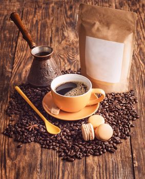 yellow cup of coffee, macaroons, beans, turkish coffee pot, and craft paper pouch bag on wooden backgroun