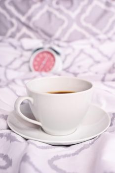 Close up of a coffee cup placed on modern bed sheets with an alarm clock in the background. Morning coffee in bed concept. Morning coffee in bed. White and grey textured bedding.