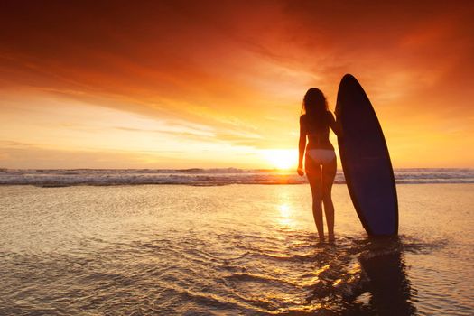 Silhouette of woman on tropical beach holding surfboard at sunset in Bali