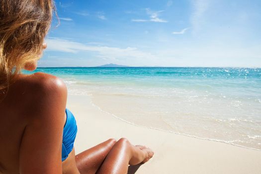 Beautiful tan woman in bikini lies on white sand beach looking on the sea, back view