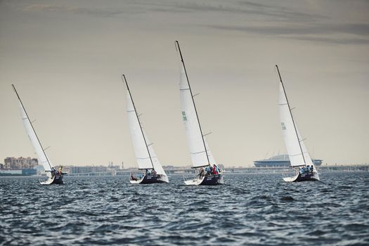 Russia, St.Petersburg, 05 September 2020: Some sailboats in a list goes by sea, the storm sky, regatta, Gazprom arena stadium on background. High quality photo