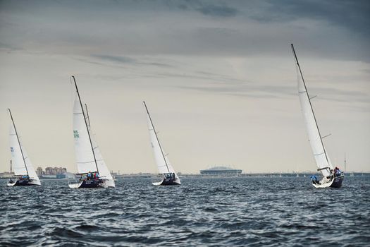 Russia, St.Petersburg, 05 September 2020: Some sailboats in a list goes by sea, the storm sky, regatta, Gazprom arena stadium on background. High quality photo