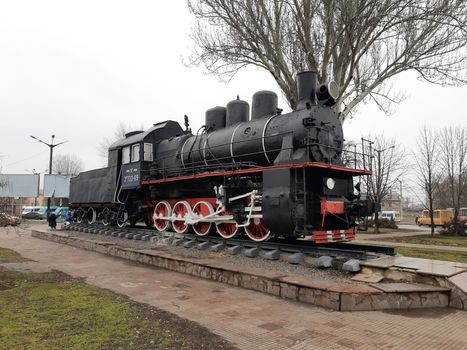 An old black steam locomotive on rails and a concrete platform in the middle of a city square.