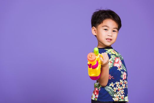 Happy Asian little boy holding plastic water gun, Thai kid funny hold toy water pistol and smiling, studio shot isolated on purple background, Thailand Songkran festival day national culture party