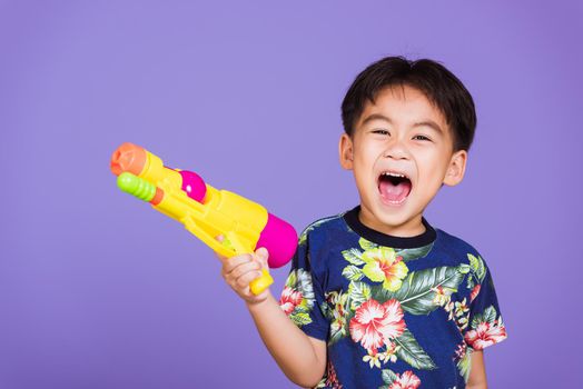 Happy Asian little boy holding plastic water gun, Thai kid funny hold toy water pistol and smiling, studio shot isolated on purple background, Thailand Songkran festival day national culture party