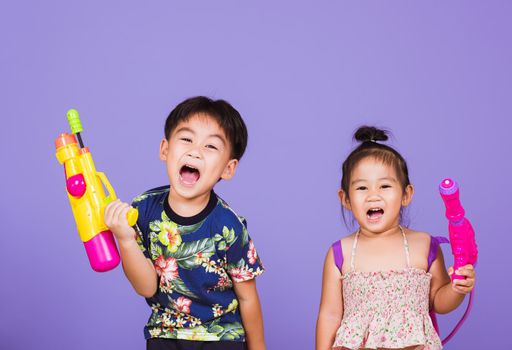 Two Happy Asian little boy and girl holding plastic water gun, Thai children funny hold toy water pistol and smile, studio shot isolated on purple background, Thailand Songkran festival day culture.