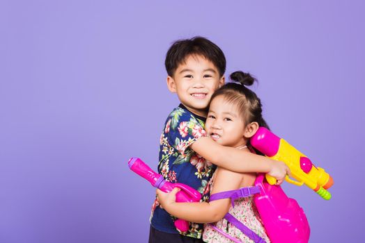 Two Happy Asian little boy and girl holding plastic water gun, Thai children funny hold toy water pistol and smile, studio shot isolated on purple background, Thailand Songkran festival day culture.