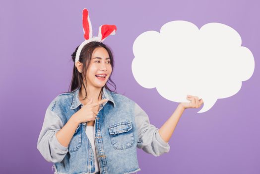 Happy Easter Day. Beautiful young woman smiling excited wearing rabbit ears and denims holding empty speech bubble, Portrait female looking at bubbles, studio shot isolated on purple background