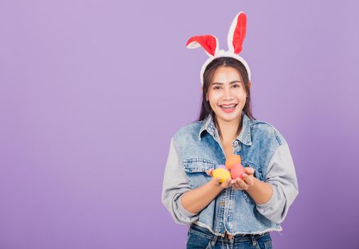 Happy Easter concept. Beautiful young woman smiling wearing rabbit ears and denims hold colorful Easter eggs gift on hands, Portrait female looking at camera, studio shot isolated on purple background