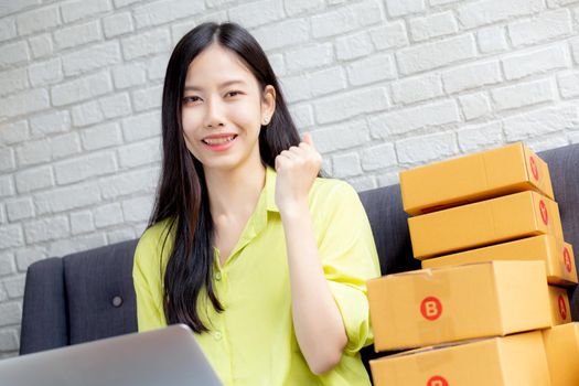 Young asian woman is merchant using laptop computer on sofa checking order of customer with purchase online shopping, business SME or startup, entrepreneur or seller and service delivery product.