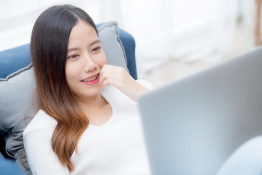Young asian business woman smile and work from home with laptop computer online to internet on sofa in living room, freelance girl using notebook on couch with comfort, new normal, lifestyle concept.