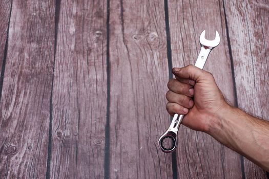 combination wrench in a fist on a wooden background. chrome-plated shiny tool with ratchet handle. The hand is dirty.