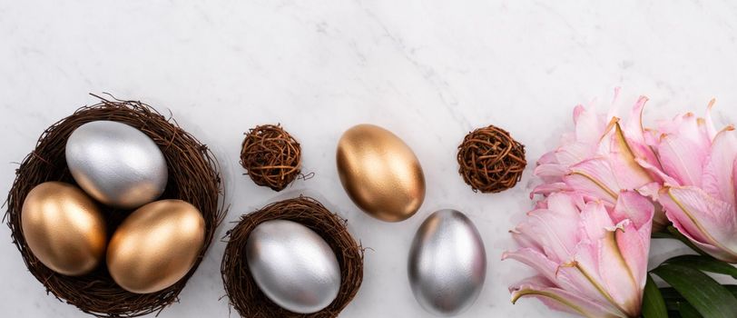 Design concept of Golden and silver Easter eggs in the nest with pink lily flower on bright marble white table background.