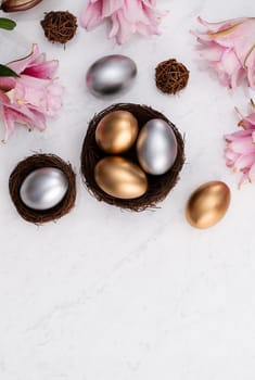 Design concept of Golden and silver Easter eggs in the nest with pink lily flower on bright marble white table background.