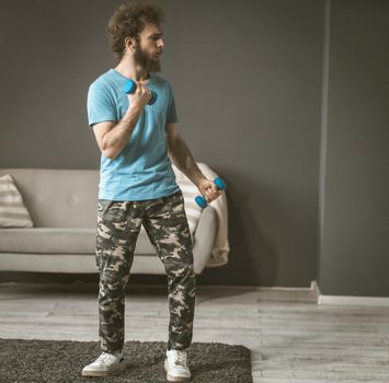 A young man sports in his room. Exercise with dumbbells. The curly-haired guy is holding dumbbells in his hands. High quality photo