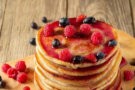 Stack of pancakes with syrup and fresh berries on wooden board on table.