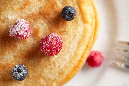 Stack of pancakes with fresh berries on a white plate close up, top view.