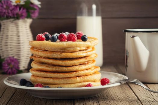 Stack of pancakes with fresh berries on a white plate, a bottle of milk and a vase of wildflowers on the table.