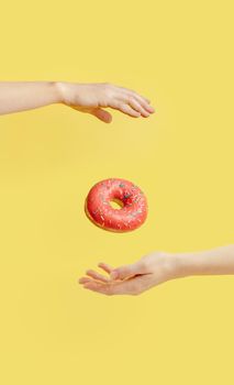 Donut falls between hands on bright yellow paper background. Donuts delivery, diet, sugar concept. Female hands and glazed donut with sprinkles flying in air. Vertical. Creative still life