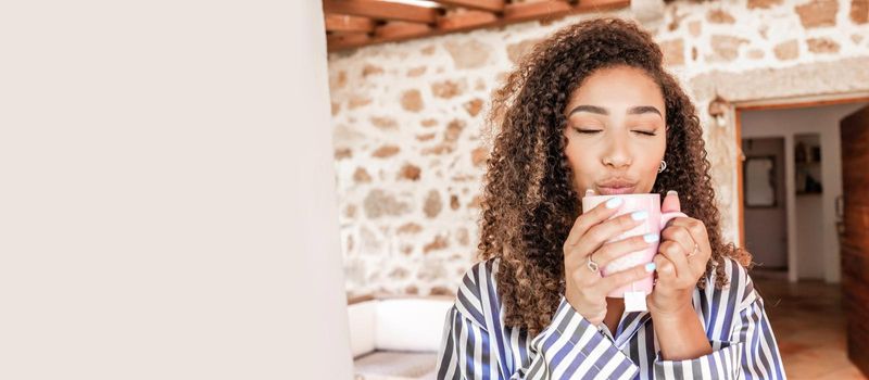 Beautiful black Hispanic girl with dark curly hair in striped silk pajamas enjoying her cup of hot tea holding it in her hands with her eyes closed on the porch of her country house. Left copy space