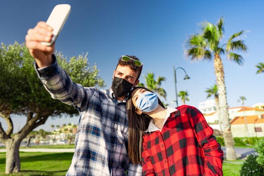 Young student couple in checkered shirt takes selfie wearing protective mask due to Coronavirus pandemic. Young Caucasian man photographs himself with girlfriend leaning on his shoulder affectionately