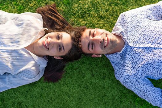 Young couple of beautiful people lying on the ground on a green lawn at sunset or sunrise head to head looking at camera from below laughing happily and confidently in the future. Concept of wellbeing