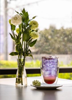 Glass of soda drink with lemonade, orange mixer butterfly pea juice (Anchan) cold refreshing drink or beverage with ice and A bouquet of white roses in a glass vase on white table. Selective focus.